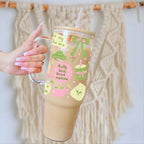 A hand with pink manicured nails holds a clear handled tumbler with a straw, filled with a light brown iced drink. The tumbler has a bamboo lid and a cute pastel graphic featuring matcha, green bows, and the text 'Pretty Girls Drink MATCHA'.