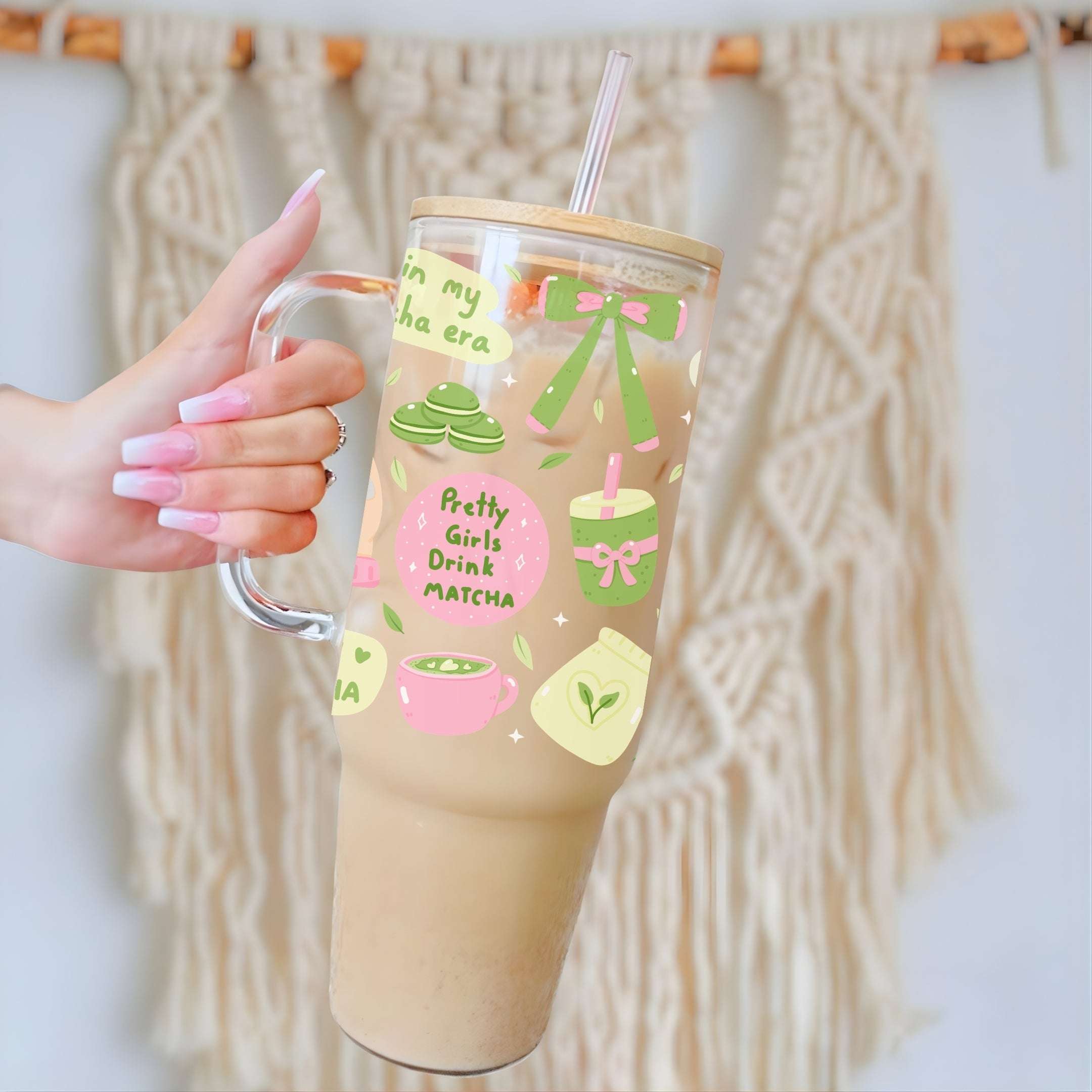 A hand with pink manicured nails holds a clear handled tumbler with a straw, filled with a light brown iced drink. The tumbler has a bamboo lid and a cute pastel graphic featuring matcha, green bows, and the text 'Pretty Girls Drink MATCHA'.