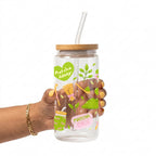 A hand holding a clear glass can-shaped tumbler with a bamboo lid and clear straw. The tumbler is decorated with green and pink illustrations of matcha leaves, a matcha bag, and the text 'Matcha Lover' and 'Matcha Girlie'.