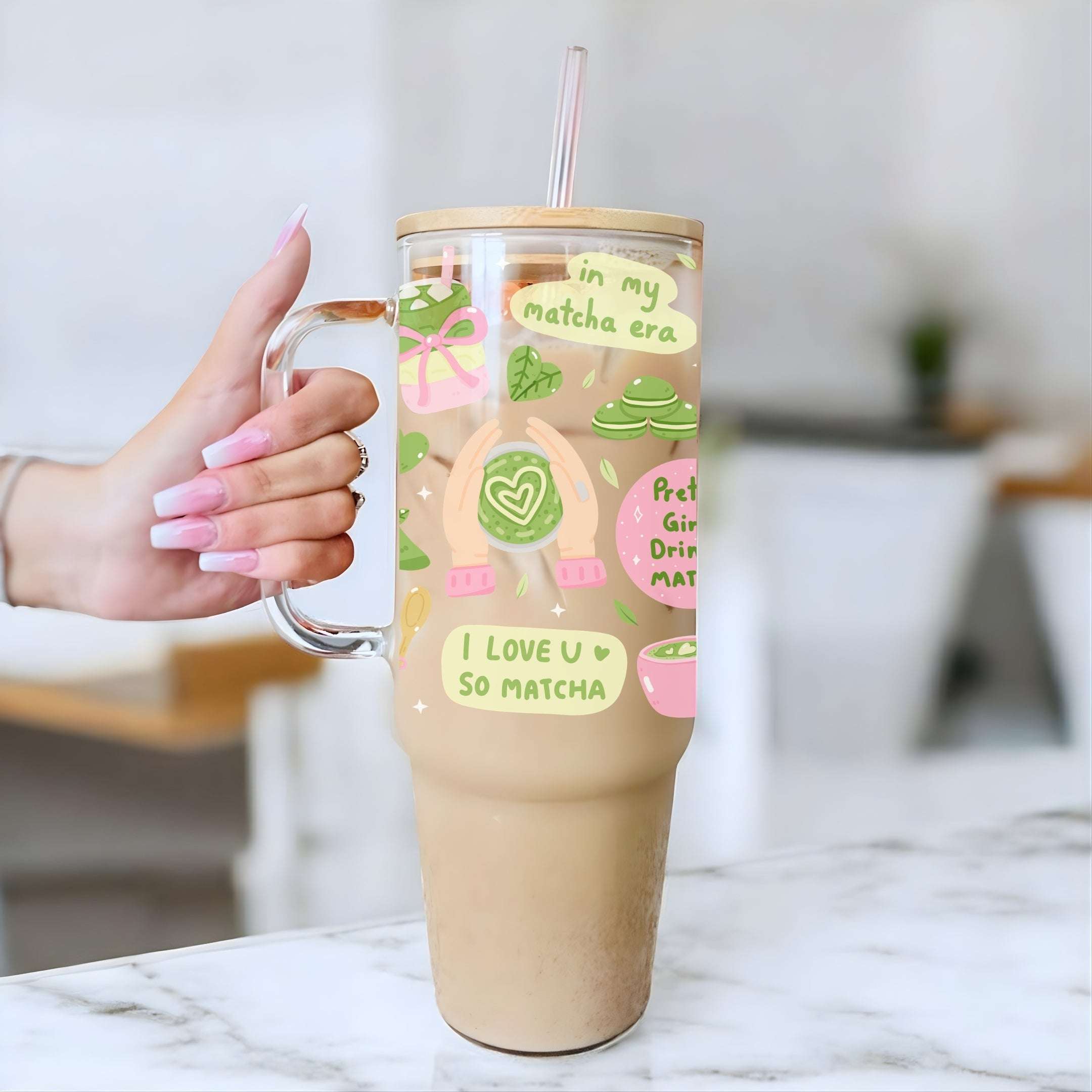 A hand with pink manicured nails holds a clear handled tumbler with a straw, filled with a light brown matcha latte. The tumbler features cute pastel illustrations of matcha green tea cakes, sweets, and text that reads 'I LOVE U SO MATCHA'.