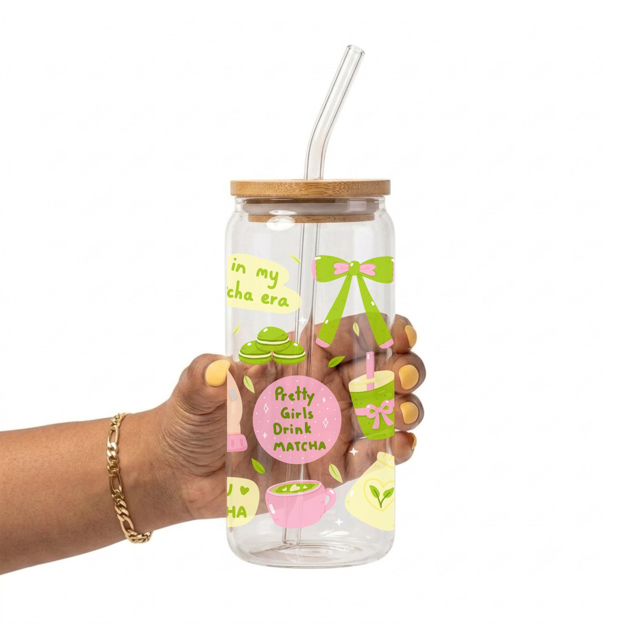 A hand holding a clear glass water bottle with a bamboo lid and clear straw. The bottle features colorful illustrations of matcha green tea, pink desserts, and a pink circle that reads 'Pretty Girls Drink MATCHA'.