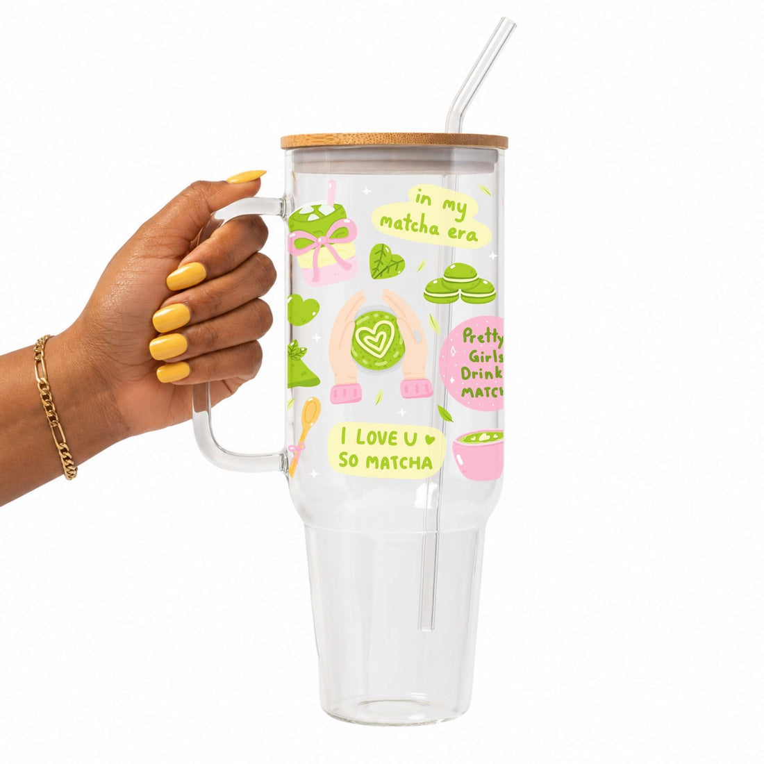 A hand with yellow nail polish holds a clear handled tumbler with a bamboo lid and straw. The tumbler is decorated with green and pink matcha-themed illustrations and text including 'in my matcha era' and 'I LOVE U SO MATCHA'.