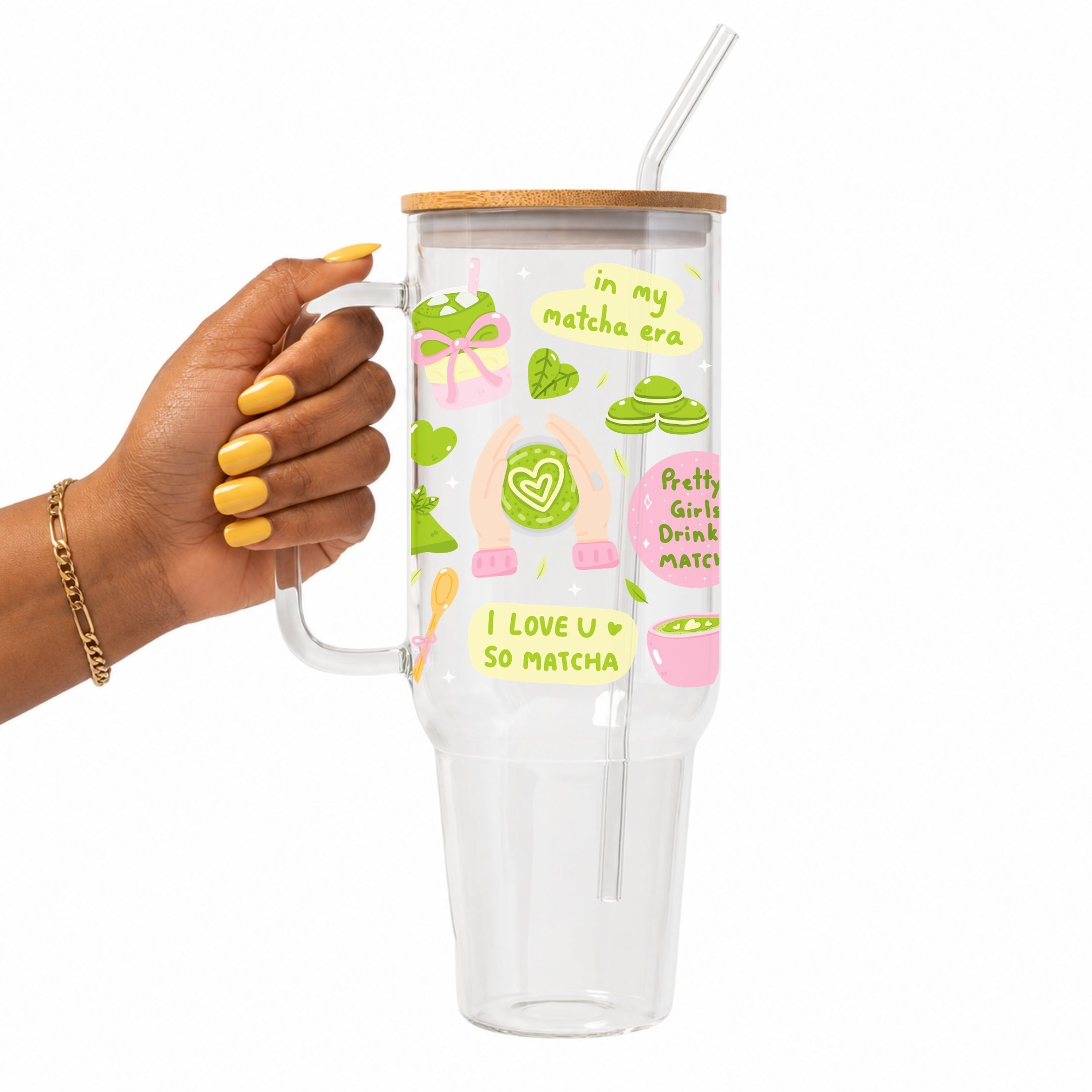 A hand with yellow nail polish holds a clear handled tumbler with a bamboo lid and straw. The tumbler is decorated with green and pink matcha-themed illustrations and text including 'in my matcha era' and 'I LOVE U SO MATCHA'.
