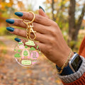 A person's hand with teal nail polish holds a round clear acrylic keychain featuring a matcha-themed illustration with green and pink details and gold hardware, set against an outdoor autumn background.