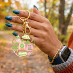 A person's hand with teal nail polish holds a round clear acrylic keychain featuring a matcha-themed illustration with green and pink details and gold hardware, set against an outdoor autumn background.