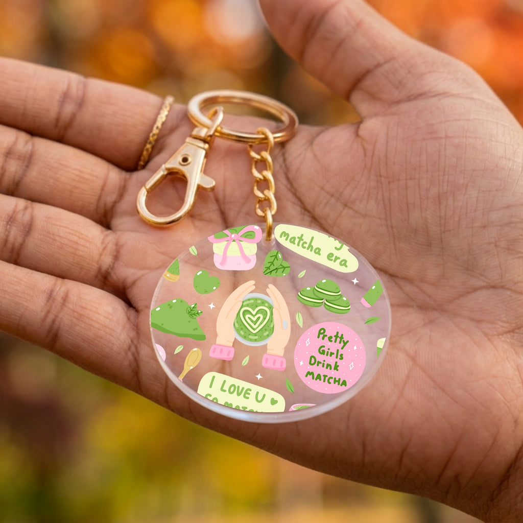 A round acrylic keychain charm held in a person's hand, featuring colorful illustrations of matcha green tea elements and text including 'Matcha Era' and 'Pretty Girls Drink Matcha'. The keychain has a gold-tone clasp and chain.