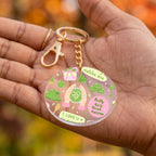 A round acrylic keychain charm held in a person's hand, featuring colorful illustrations of matcha green tea elements and text including 'Matcha Era' and 'Pretty Girls Drink Matcha'. The keychain has a gold-tone clasp and chain.