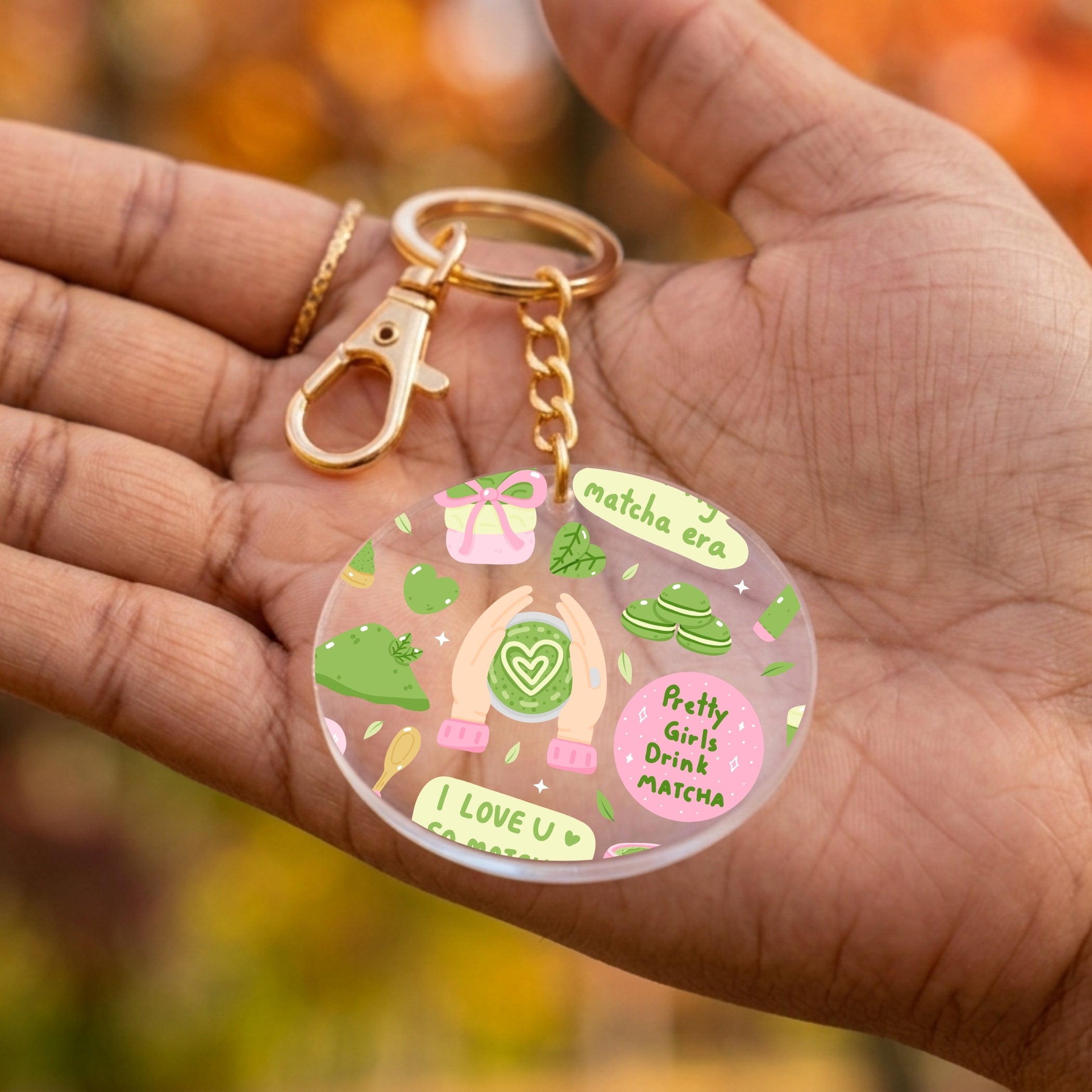 A round acrylic keychain charm held in a person's hand, featuring colorful illustrations of matcha green tea elements and text including 'Matcha Era' and 'Pretty Girls Drink Matcha'. The keychain has a gold-tone clasp and chain.