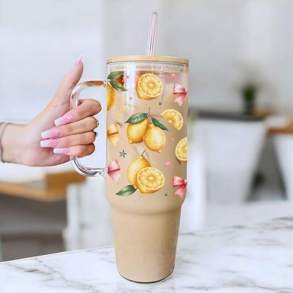 A hand with pink French-tipped nails holds a clear handled tumbler filled with a light brown iced coffee or latte. The tumbler features a printed pattern of yellow lemons, green leaves, and pink bows, with a clear lid and straw.