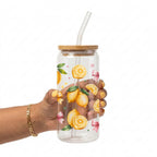 A hand holding a clear glass water bottle with a bamboo lid and a clear bent straw. The bottle is decorated with illustrations of lemons, pink bows, and green leaves.