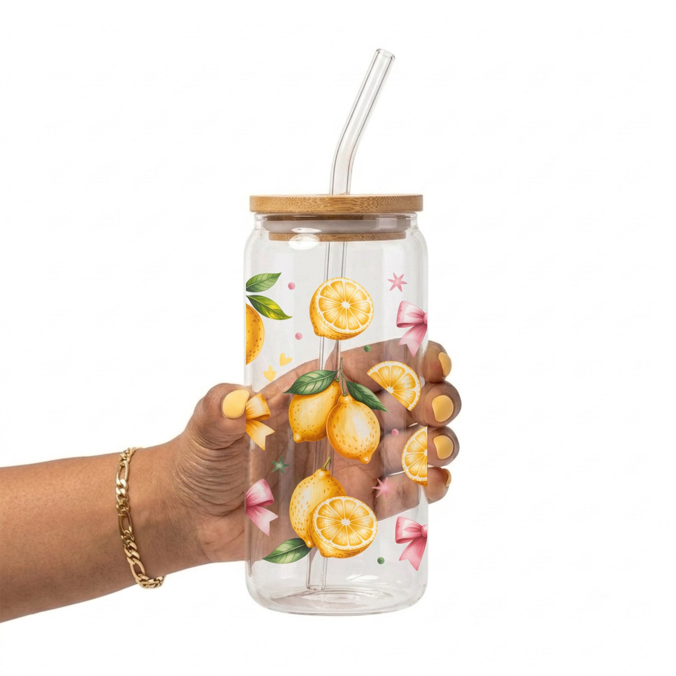 A hand holding a clear glass water bottle with a bamboo lid and a clear bent straw. The bottle is decorated with illustrations of lemons, pink bows, and green leaves.