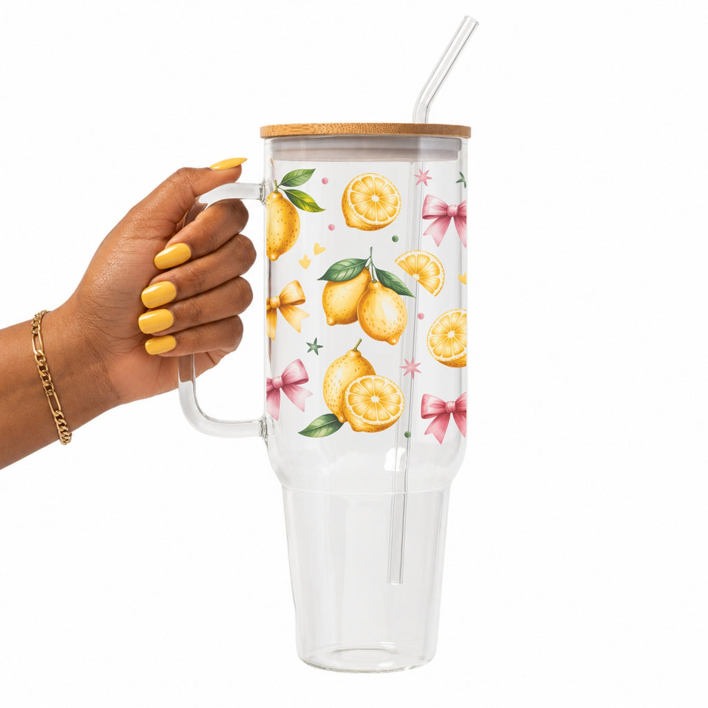 A hand with yellow nails holds a clear double-wall tumbler with a bamboo lid and straw. The tumbler features a white printed pattern of lemons, green leaves, and pink bows.