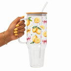 A hand with yellow nails holds a clear double-wall tumbler with a bamboo lid and straw. The tumbler features a white printed pattern of lemons, green leaves, and pink bows.