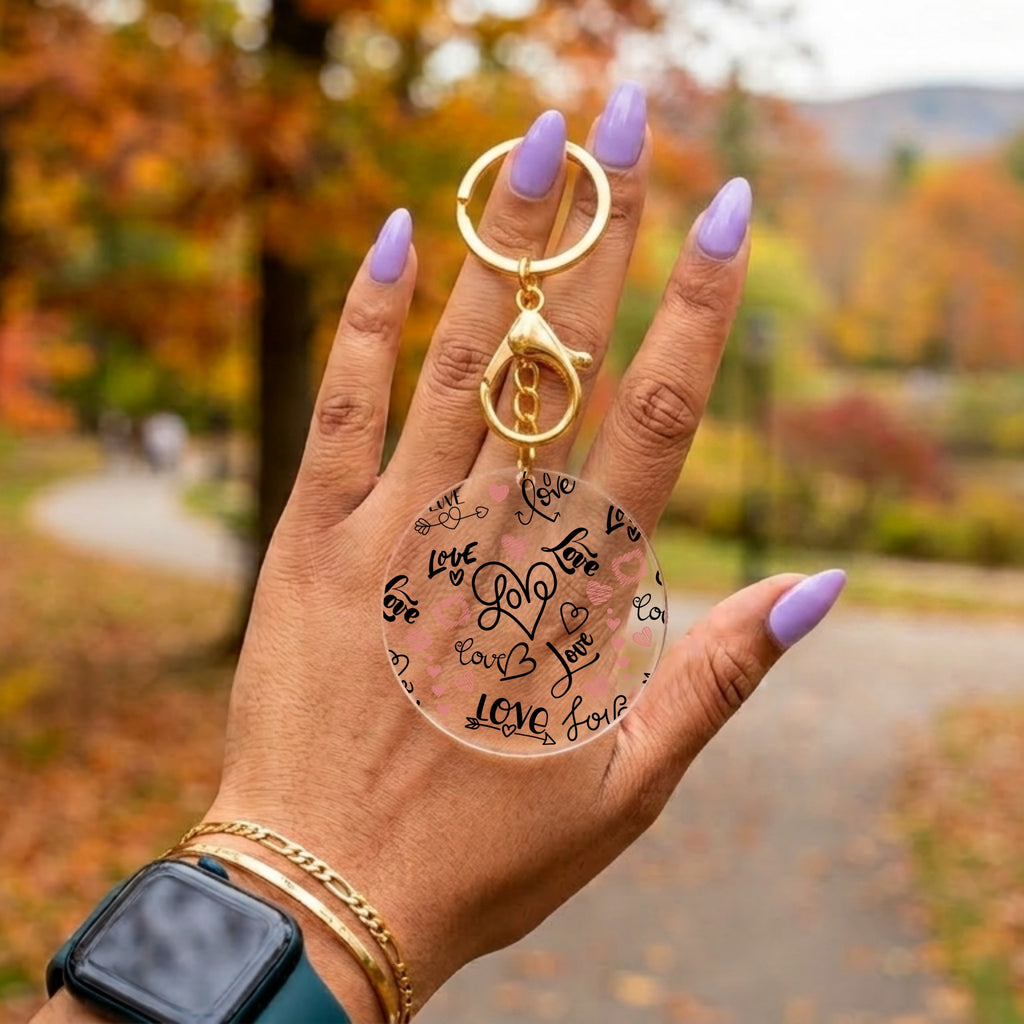 A hand with lavender nail polish holds a clear acrylic keychain with black script writing and gold-tone hardware against a blurred autumn park background.