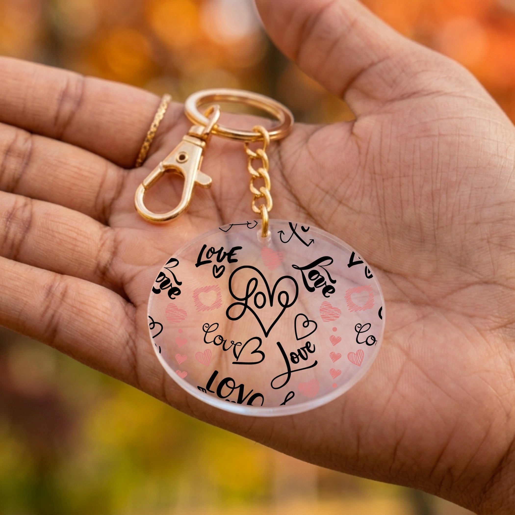 A round acrylic keychain with a gold clasp and chain, held in a person's hand. The keychain features a frosted pink background with black hand-drawn hearts, the word 'Love' in script, and small pink hearts.