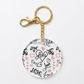 A round acrylic keychain charm featuring a white background with black and pink hand-drawn hearts, hearts, and the word 'Love' in various script styles, attached to a gold-tone split ring and clasp.