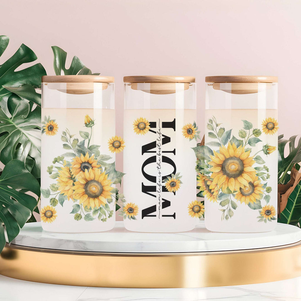 Three clear glass canisters with gold lids are arranged on a white marble tray with a gold base. The center canister has large black vertical text reading 'MOM' with small script text above and below, while the left and right canisters feature yellow sunflower and green leaf floral prints.