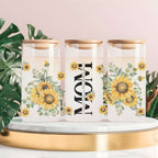 Three clear glass canisters with gold lids are arranged on a white marble tray with a gold base. The center canister has large black vertical text reading 'MOM' with small script text above and below, while the left and right canisters feature yellow sunflower and green leaf floral prints.