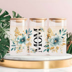Three clear glass canisters with gold-toned lids are arranged on a white marble tray with a gold rim. The center canister has the word “MOM” printed vertically in black, with smaller script text between the letters.