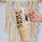 Nik's hand holding a clear tumbler with floral design and 'MOM' text against a textured wall.