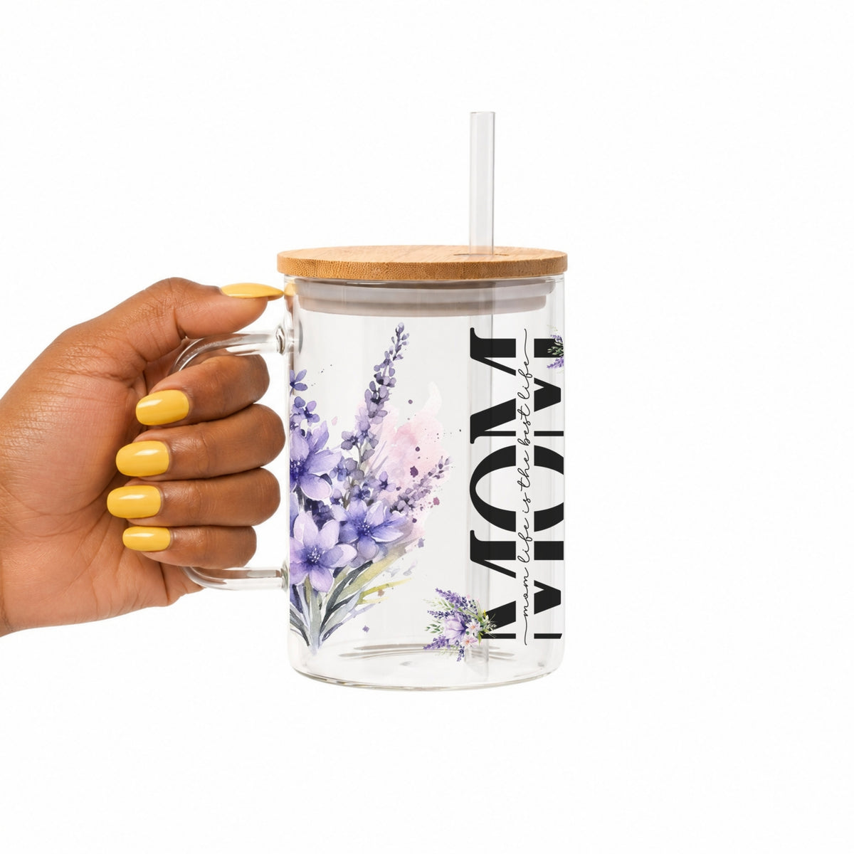 A hand with yellow nails holds a clear glass mug with a bamboo lid and straw. The mug features a purple and pink watercolor floral design and the word 'MOM' printed vertically in black.