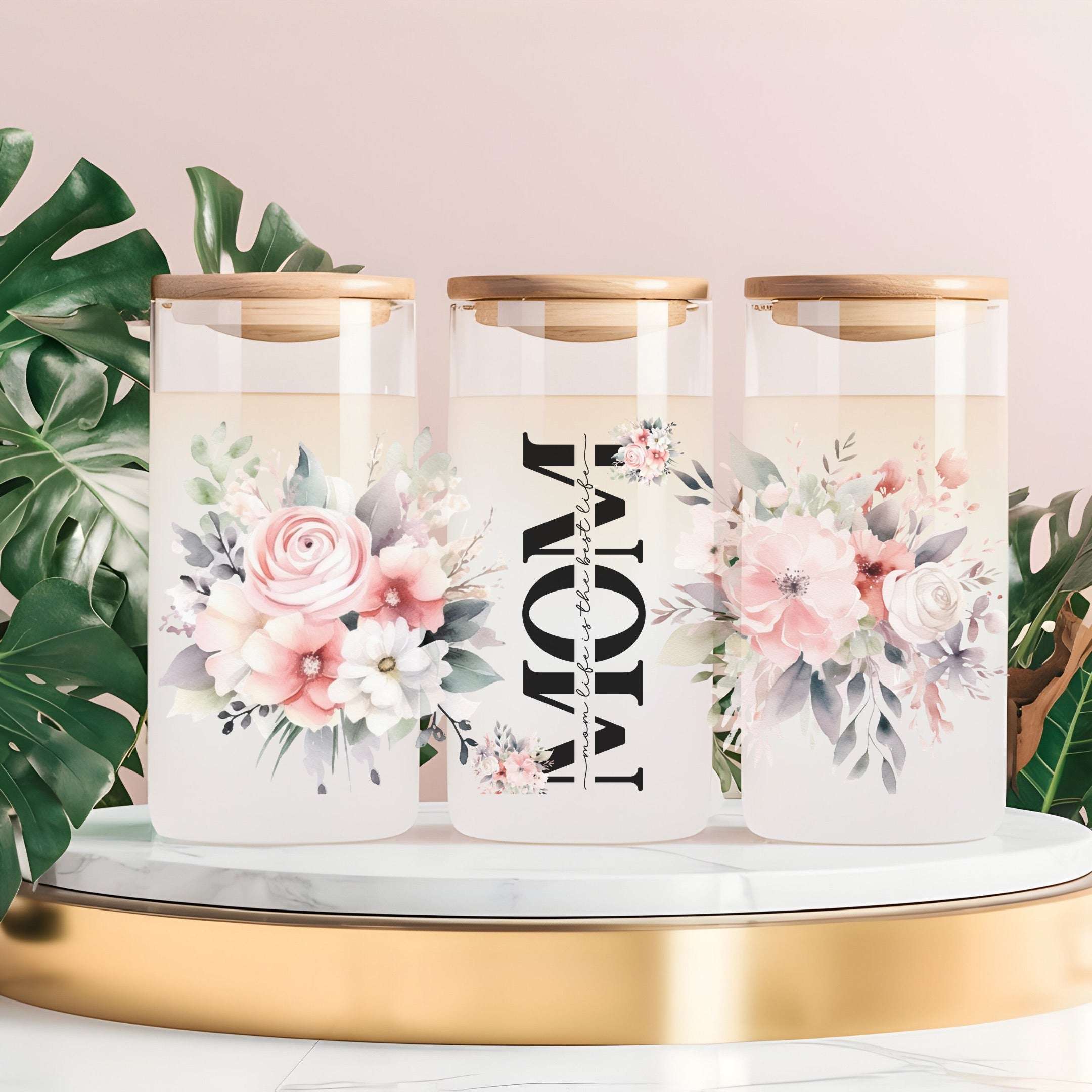 Three clear glass canisters with gold lids are arranged on a white marble-and-gold tray against a soft pink background, with green leaves at the sides. The center canister is printed with the word “MOM” vertically in black, flanked by pink and white floral illustrations.
