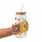 A hand holding a clear glass tumbler with a bamboo lid and clear glass straw, decorated with a yellow sunflower and green leaf pattern, against a white background.