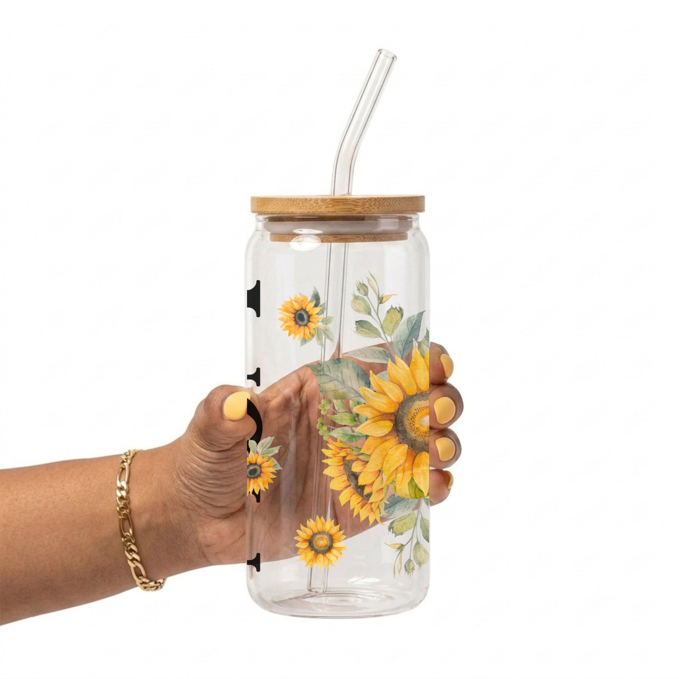 A hand holding a clear glass tumbler with a bamboo lid and clear glass straw, decorated with a yellow sunflower and green leaf pattern, against a white background.