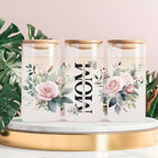 Three tall clear glass canisters with gold lids are arranged on a white marble-and-gold tray against a soft pink background, with green monstera leaves around them. The center canister has large black vertical text reading “MOM,” and the side canisters have pink rose and floral illustrations.