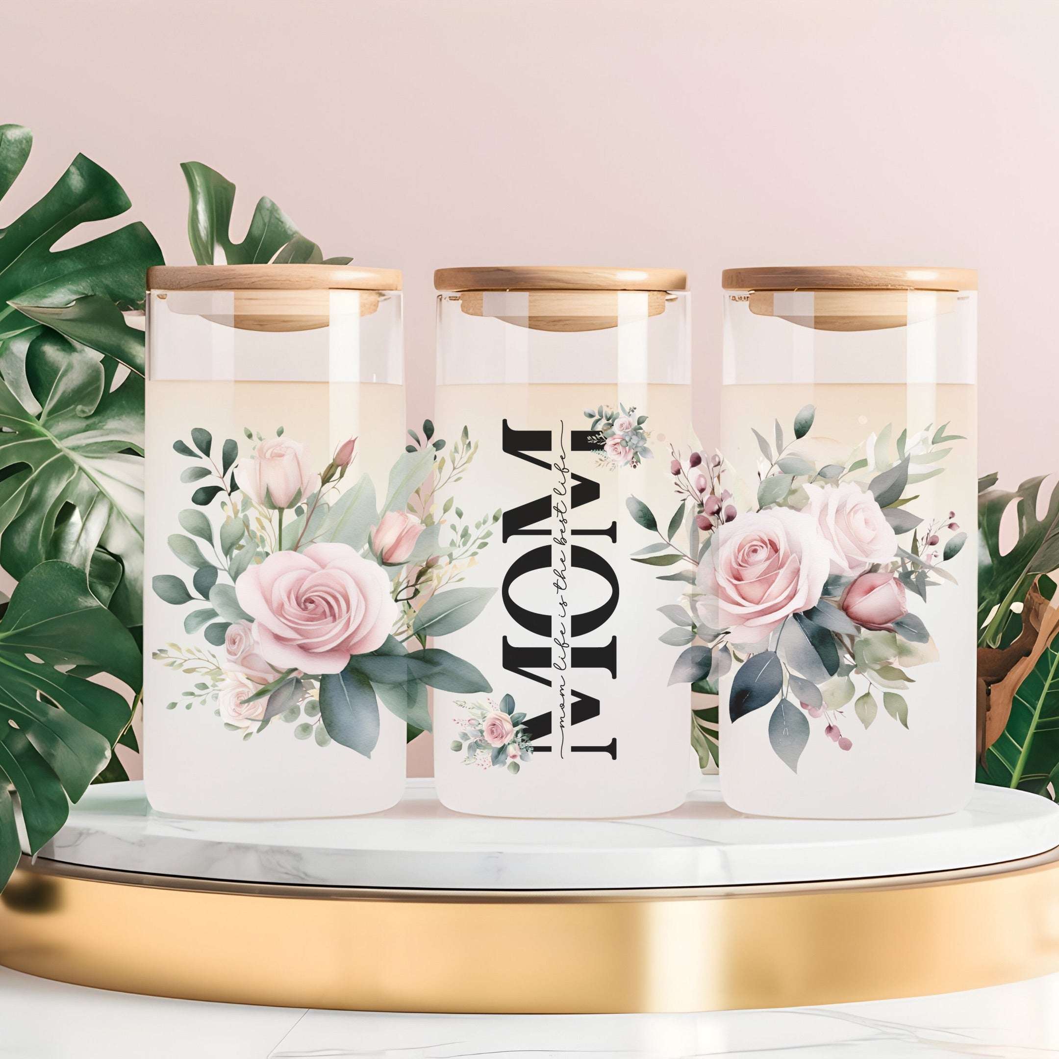 Three tall clear glass canisters with gold lids are arranged on a white marble-and-gold tray against a soft pink background, with green monstera leaves around them. The center canister has large black vertical text reading “MOM,” and the side canisters have pink rose and floral illustrations.