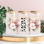 Three clear glass canisters with gold lids are arranged on a white marble-and-gold pedestal against a soft pink background, with green monstera leaves around them. The center canister has large black text reading “MOM” with smaller script text between the words, and the side canisters have pink and white floral prints.