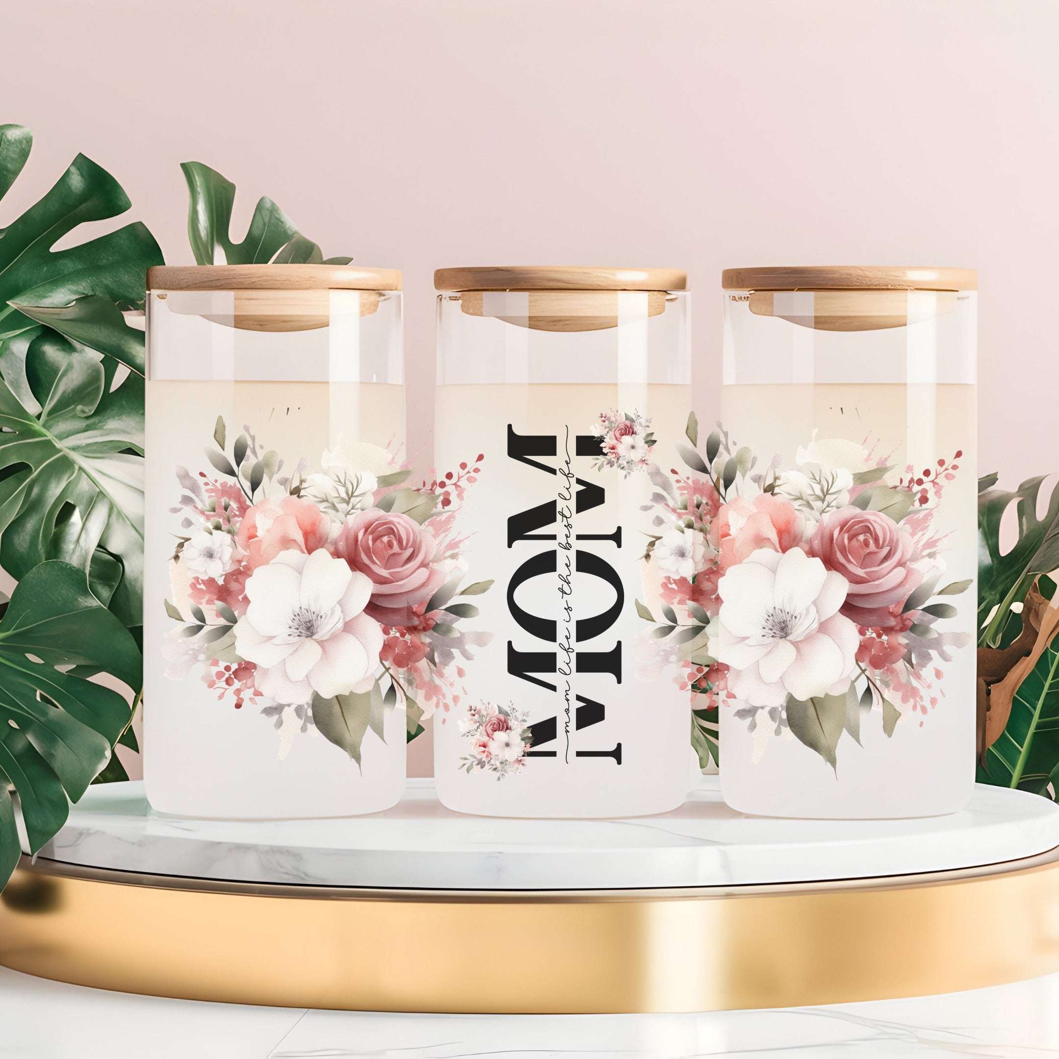 Three clear glass canisters with gold lids are arranged on a white marble-and-gold pedestal against a soft pink background, with green monstera leaves around them. The center canister has large black text reading “MOM” with smaller script text between the words, and the side canisters have pink and white floral prints.