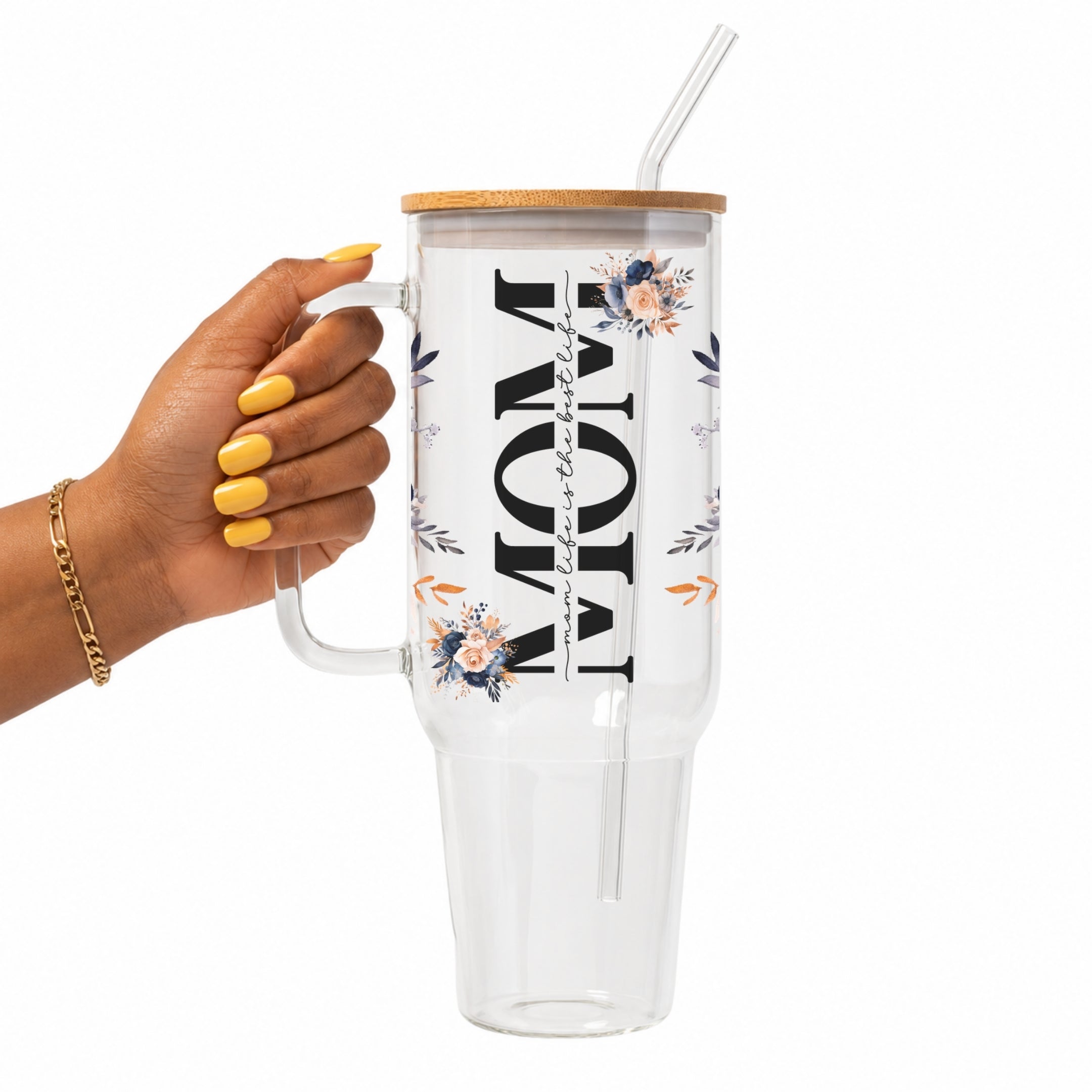A hand with yellow nails holds a clear handled tumbler with a bamboo lid and clear straw. The tumbler features a floral design and the word 'MOM' printed vertically in black, with 'with love' written in a smaller script.