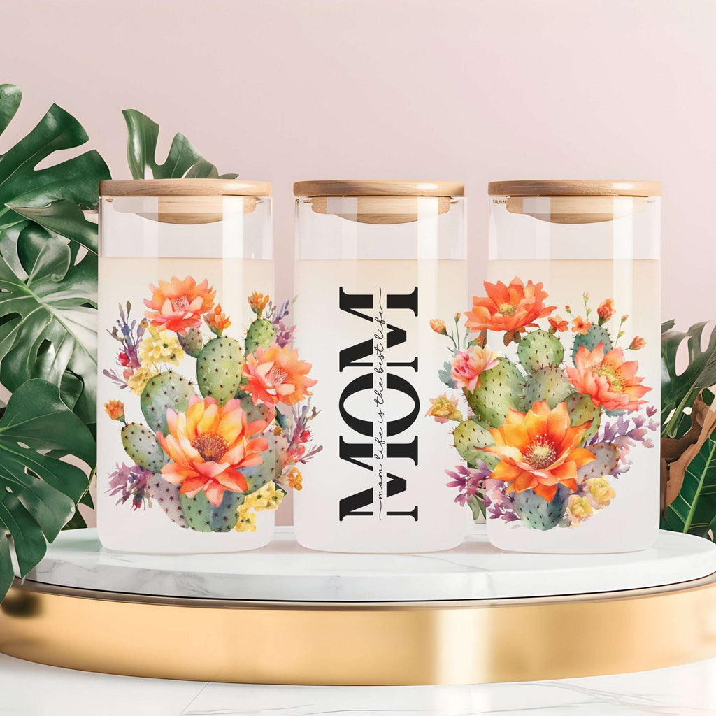 Three clear glass canisters with light wood lids are arranged on a gold-rimmed white tray. The center canister has the word “MOM” printed vertically in black, with smaller text that appears to say “with love,” while the side canisters feature colorful cactus and flower illustrations.