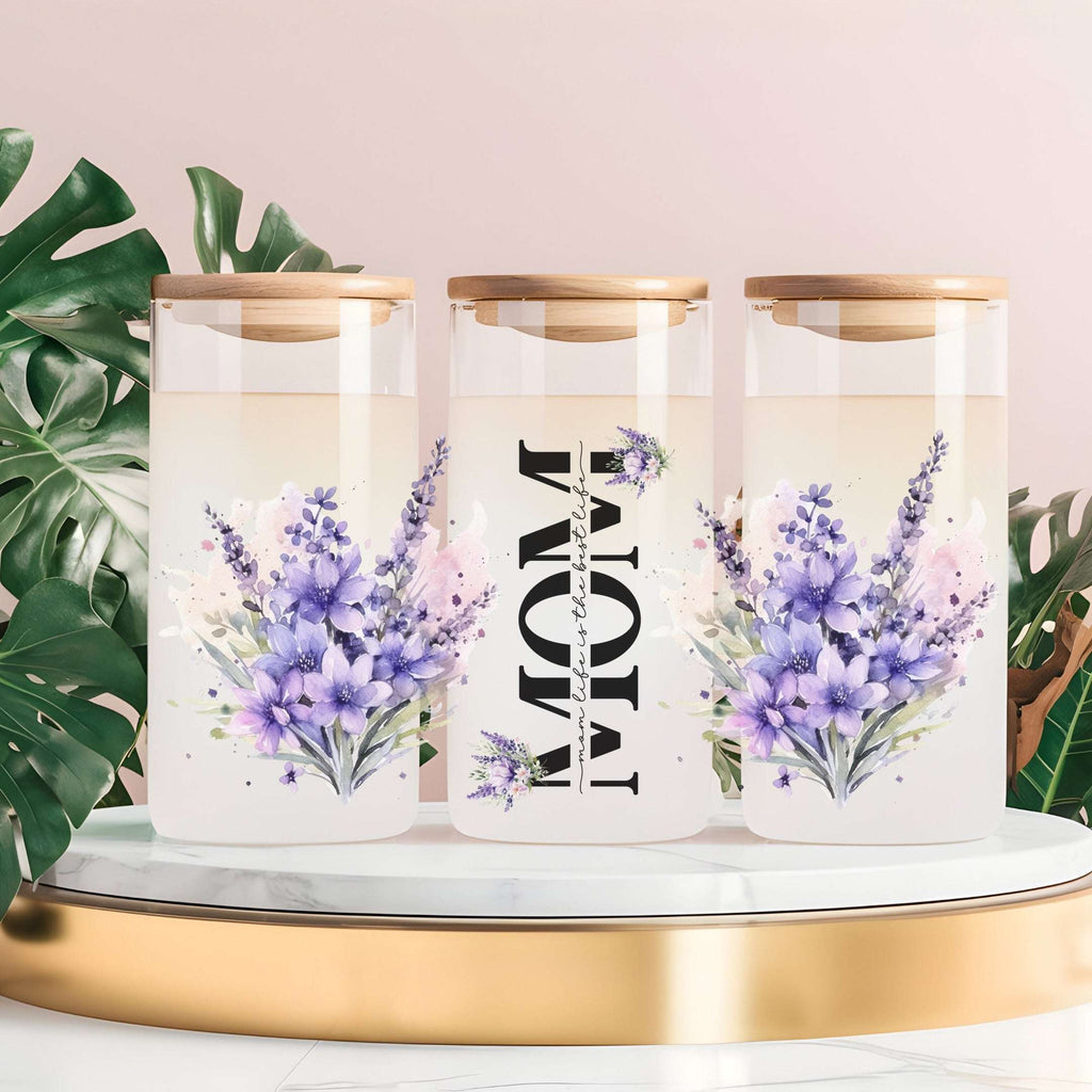 Three clear glass canisters with gold lids are arranged on a white marble-and-gold pedestal. The center canister is labeled 'MOM' vertically, and all three feature purple and pink floral watercolor designs.