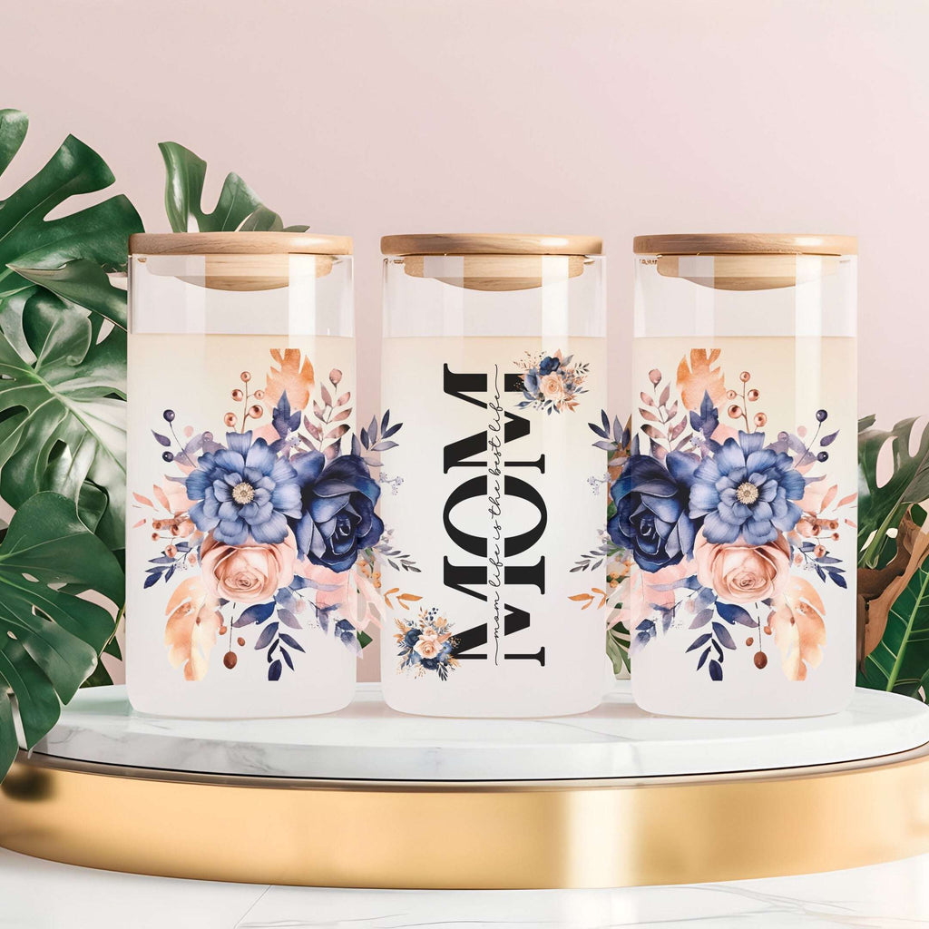 Three tall clear glass canisters with gold lids are arranged on a white marble tray with a gold base. The center canister has large black text reading 'MOM' with 'with love' in smaller script, while the left and right canisters have blue, peach, and navy floral prints.