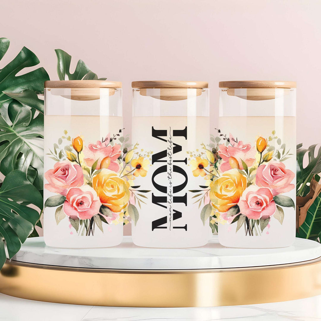 Three clear glass canisters with gold-toned lids are arranged on a white marble slab with a gold base. The center canister is labeled 'MOM' in large black letters, and the side canisters feature watercolor floral designs with pink and yellow roses, green leaves, and small yellow flowers.