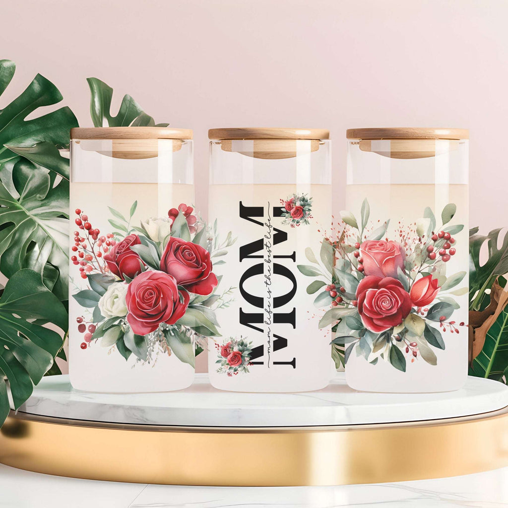 Three clear glass canisters with gold lids are arranged on a white marble-and-gold pedestal. The center canister is labeled 'MOM' vertically in black text, flanked by red rose and green leaf floral illustrations.