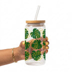 A hand holding a clear glass canister tumbler with a bamboo lid and clear glass straw, decorated with a green and yellow monstera leaf pattern, against a solid white background.