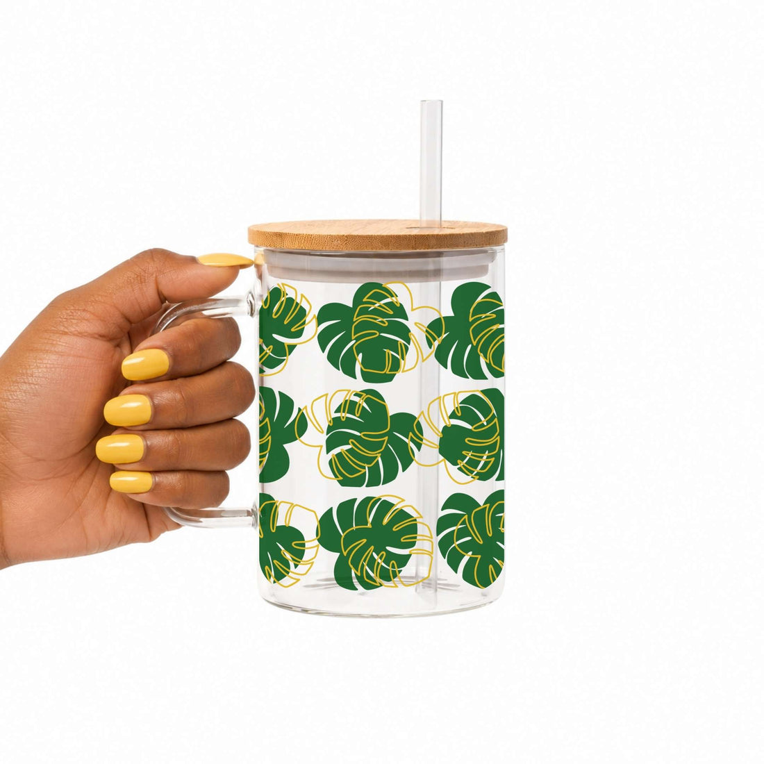 A hand with yellow nail polish holds a clear glass mug with a bamboo lid and clear straw. The mug features a pattern of green monstera leaves with gold outlines on a white background.