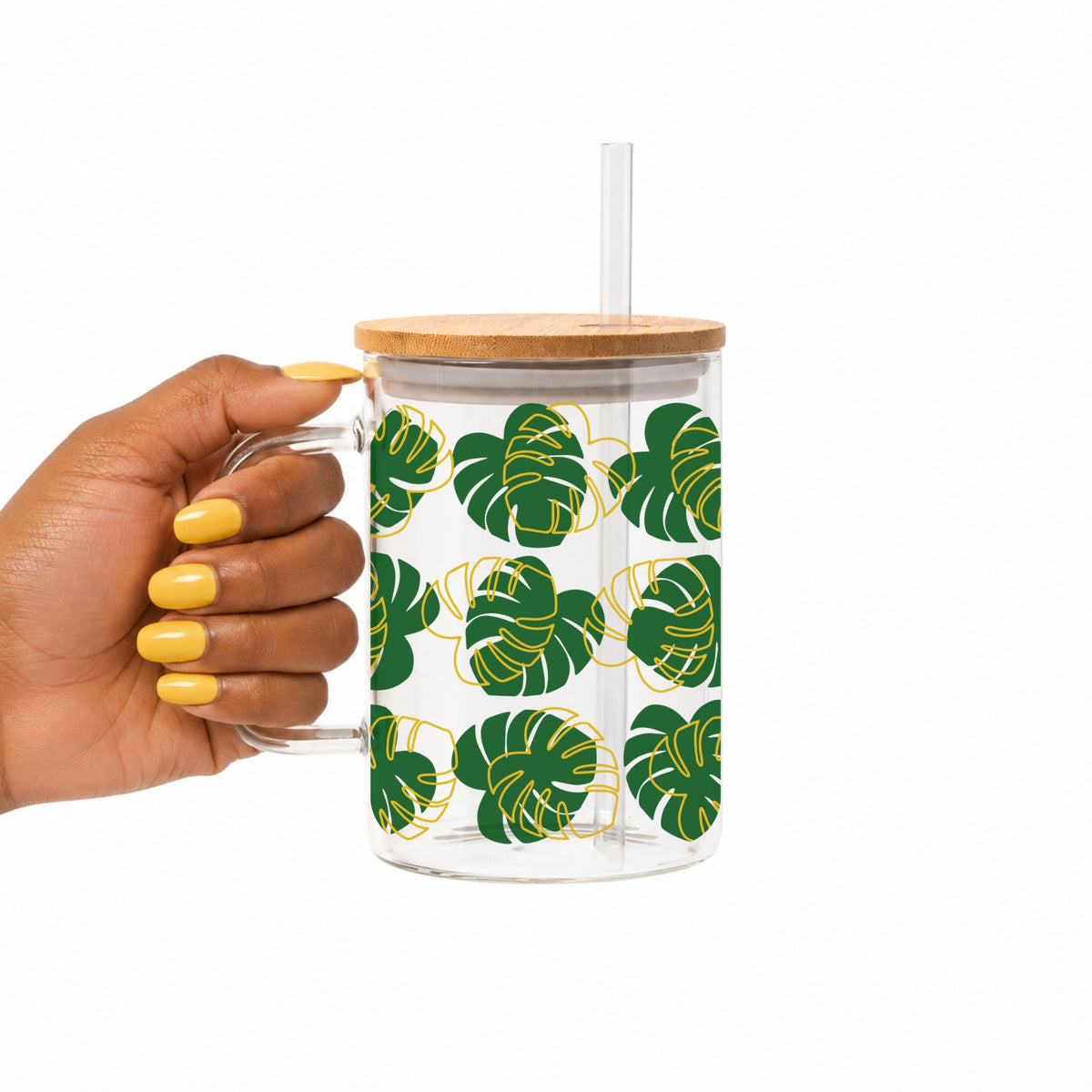 A hand with yellow nail polish holds a clear glass mug with a bamboo lid and clear straw. The mug features a pattern of green monstera leaves with gold outlines on a white background.