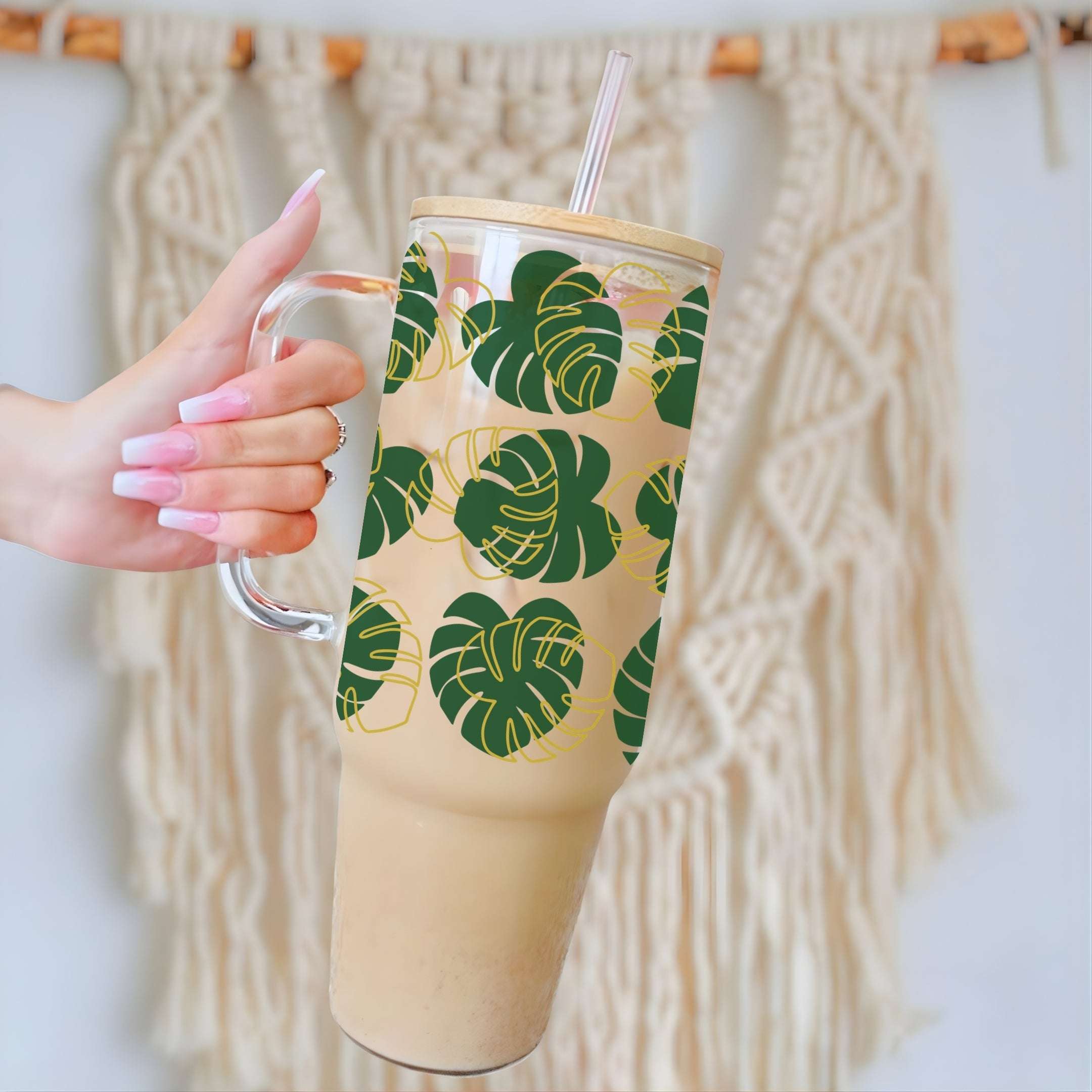 A hand with pink manicured nails holds a tall clear glass tumbler with a handle, a bamboo lid, and a clear straw. The tumbler features a pattern of green monstera leaves with gold outlines on a beige background.