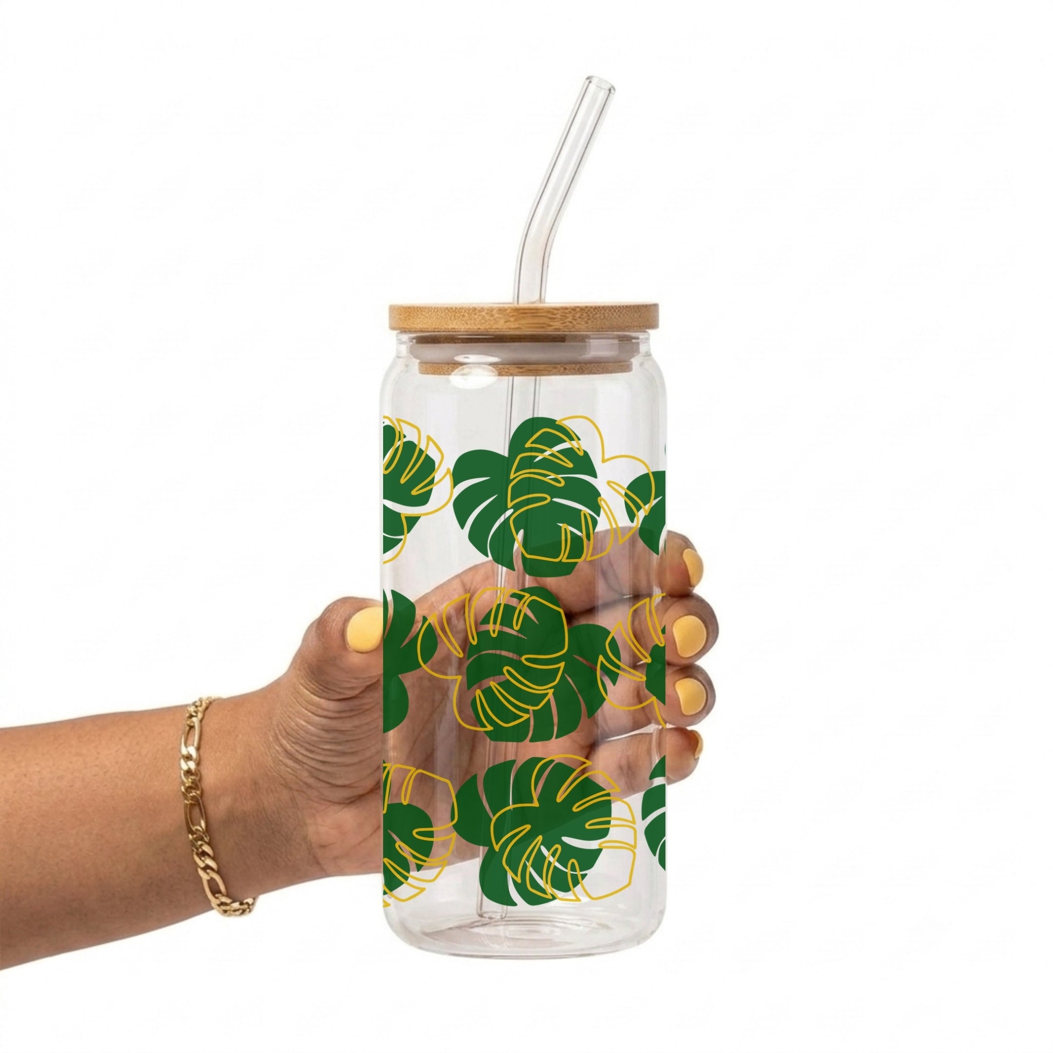 A hand holding a clear glass water bottle with a bamboo lid and clear glass straw, decorated with a green and yellow monstera leaf pattern, against a solid white background.