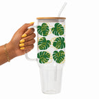 A hand with yellow nail polish holds a clear handled tumbler with a bamboo lid and clear straw. The tumbler has a white background with a green and yellow monstera leaf pattern.