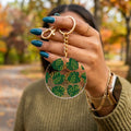 A person outdoors holds up a round clear acrylic keychain charm decorated with green tropical monstera leaves outlined in gold. The keychain has a gold split ring and chain.