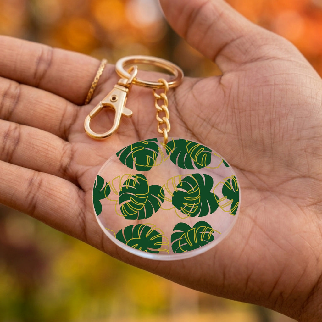 A gold-tone keychain with a round clear acrylic charm featuring a green monstera leaf pattern, held in a person's hand against a blurred autumn background.