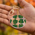 A gold-tone keychain with a round clear acrylic charm featuring a green monstera leaf pattern, held in a person's hand against a blurred autumn background.
