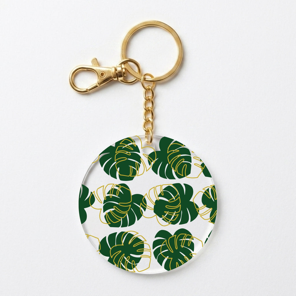 A round acrylic keychain featuring a green monstera leaf pattern with gold outlines on a white background, complete with a gold-tone clasp and split ring.
