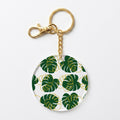 A round acrylic keychain featuring a green monstera leaf pattern with gold outlines on a white background, complete with a gold-tone clasp and split ring.