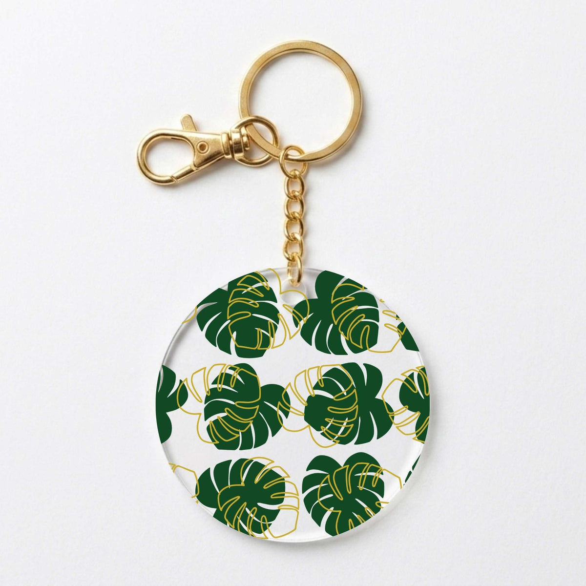 A round acrylic keychain featuring a green monstera leaf pattern with gold outlines on a white background, complete with a gold-tone clasp and split ring.